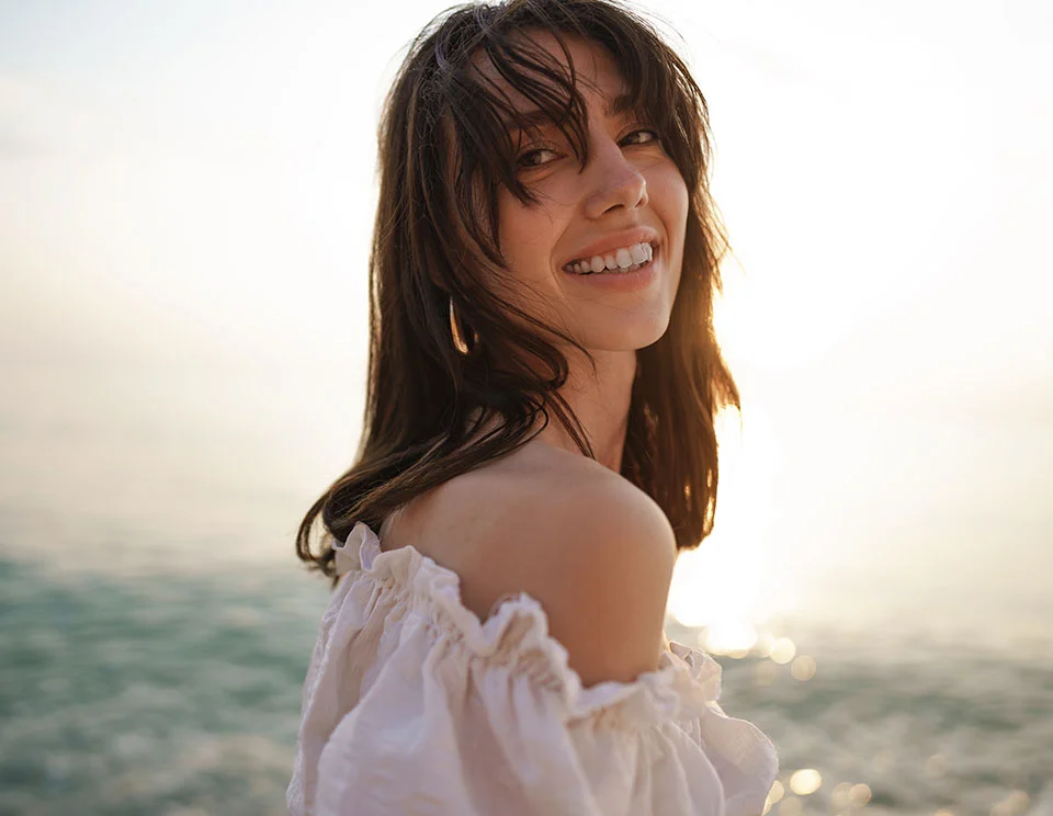 Woman standing on a beach during sunset, wearing an off-the-shoulder white top, smiling broadly as she looks back over her shoulder - Hydrafacial in Portsmouth, NH
