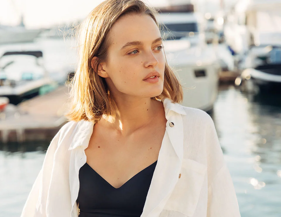 Woman standing at a marina with boats in the background, wearing a white cover-up over a black swimsuit, gazing off to the side with a relaxed expression - Laser Skin Resurfacing in Portsmouth, NH