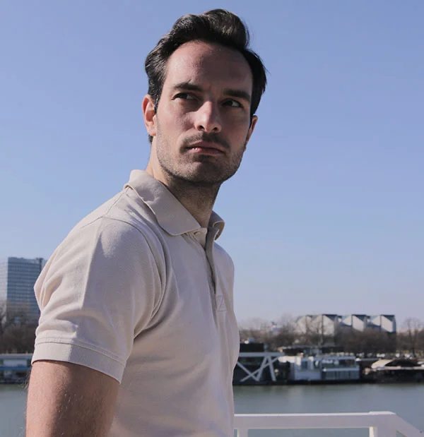 A young man with dark hair, standing outside in the sunlight, wearing a beige polo shirt. He is gazing off into the distance with a focused expression - Neck Lift in Portsmouth, NH