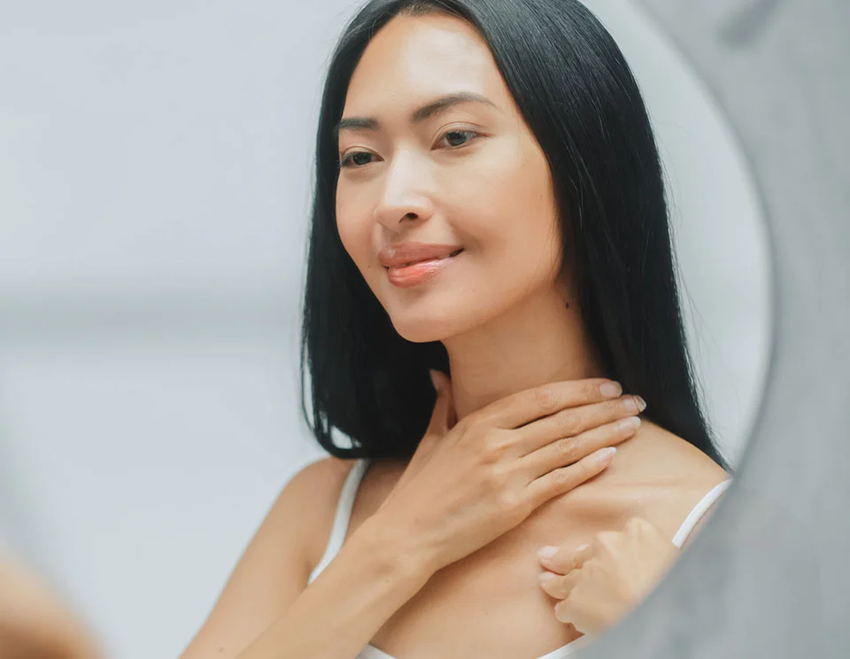 A woman with long, sleek black hair, standing in front of a mirror, gently touching her neck with a smile, appreciating her reflection - Neck Lift in Portsmouth, NH