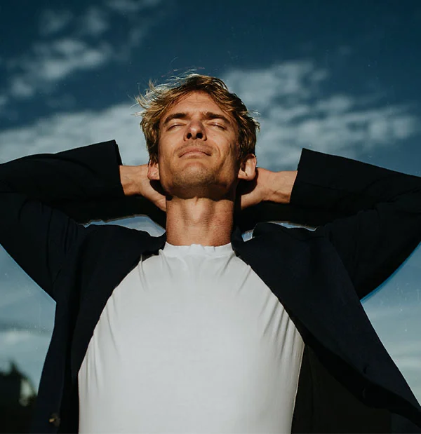 man with tousled blonde hair stands outdoors with his hands behind his head, soaking in the sunlight. He wears a dark jacket over a white shirt and has his eyes closed, embracing the peaceful moment - Ozempic Face in Portsmouth, NH